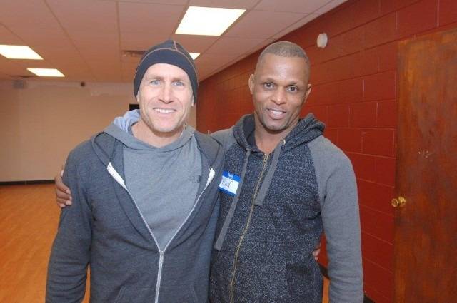 Two men smiling, arm-in-arm, wearing hoodies inside a room.